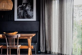 Interior of a cottage in Anglesea. Cropped photo of a dining room painted black with a timber table, a large black-and-white portrait, a rattan pendant, and sheer linen curtains opening onto bushland.