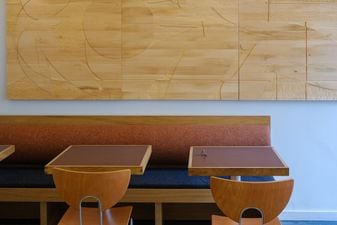 Interior of Bliskość café. Carved blond-timber panel above a rust-colored banquette seating, with oxblood tables and moulded plywood chairs.