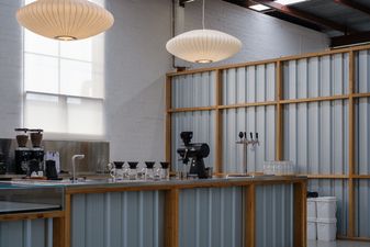 Interior of a warehouse café—Bench. Counter clad in corrugated iron and timber, with pour-over kit, grinders, and two saucer pendants over white brick.